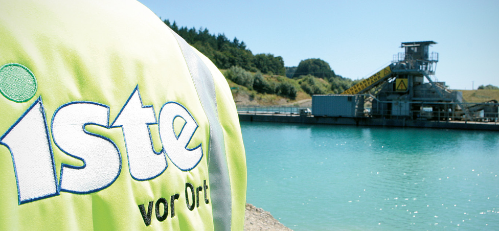 Der Rücken eines Menschen vor einem Baggersee. Auf der Weste steht 
